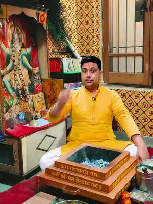 A man in a yellow shirt is sitting cross-legged on a red carpet, in front of a wooden structure containing an image of a deity. In front of him is a ritual fire pit. The background displays a golden fabric with repeating patterns. The lighting is warm and directed towards the man. Additional religious iconography and objects surround him.