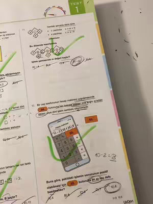 A close-up photo of a math test paper with handwritten notes and markings. The paper is white with black text and diagrams, and portions are shaded green. A calculator from a phone is shown. A hand is partially visible at the bottom of the image.