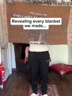 A person standing indoors, mostly obscured by a large brown blanket held up in front of them. The blanket has text, and the person is wearing black pants, a white shirt, and white slippers. The background includes furniture and decor.
