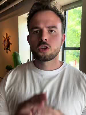 A man with short brown hair and a beard stands in front of a window, talking. He wears a white t-shirt. There's a decorative object on the wall to his left.