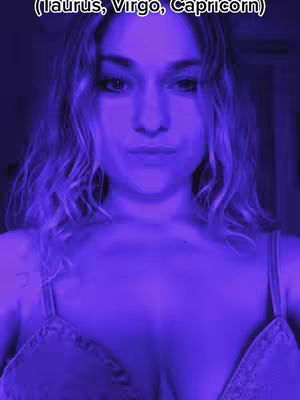 A selfie-style shot of a young woman's face and upper body. She is centered in the frame with wavy blonde hair and is wearing a strap top. The lighting has a strong, cool purple tone. The background is dimly lit.