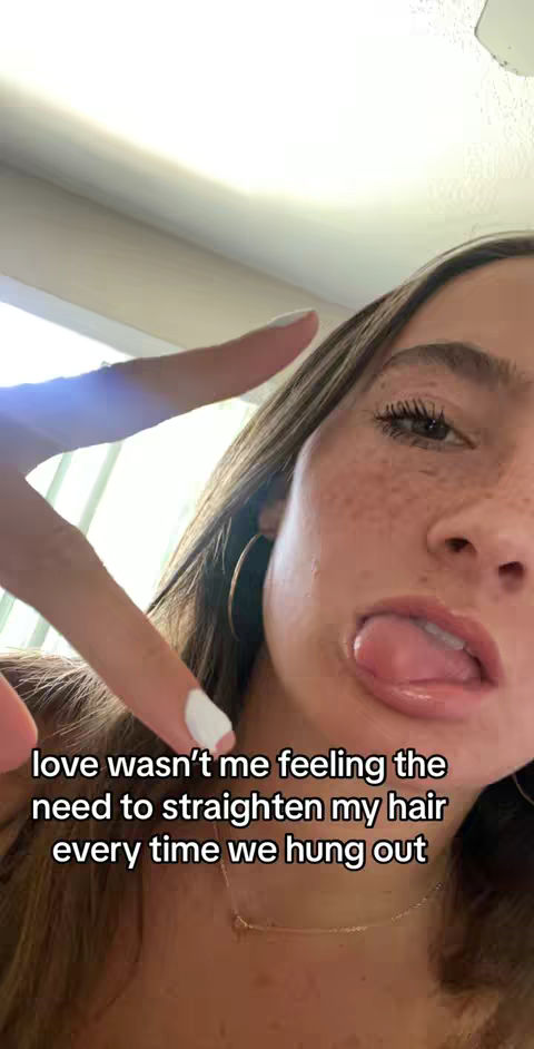 A selfie-style shot featuring a young woman with long brown hair, visible freckles, and a light-skinned complexion. She is partially making a peace sign with her fingers and sticking out her tongue. The background appears to be a generic indoor setting, possibly a room with a white ceiling and a light source.