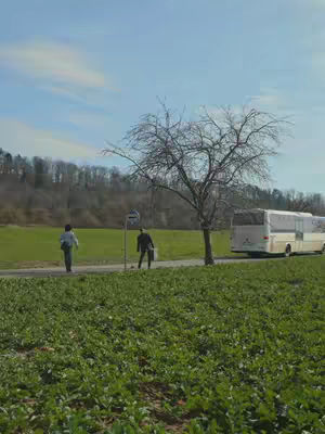 A scenic shot of a rural landscape with two people waiting at a bus stop. The sky is blue, with some clouds visible. The road is flanked on one side by a field of dense green vegetation, and on the other side by a large green field. A tree without leaves and a bus shelter are on the side of the road. A parked bus is visible in the background. The overall composition is calm and spacious.