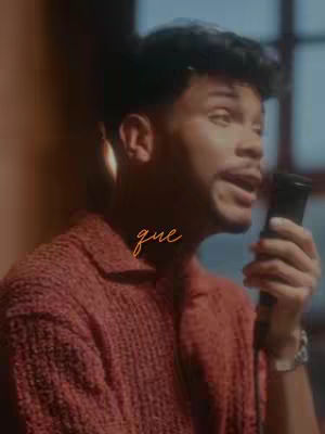 The cover image features a medium shot of a man holding a microphone and singing. The man, who appears to be of African descent, is wearing a reddish-brown knitted sweater. The background is softly lit with a blur effect that provides a sense of depth. The word 'que' is written in custom font, overlayed on the image.