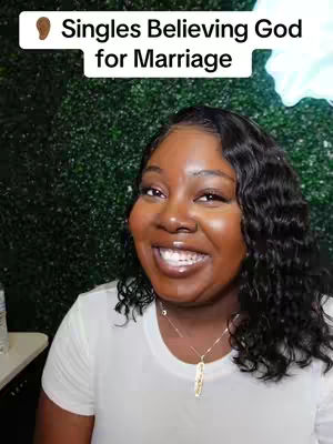 A woman with dark skin and curly black hair smiles at the camera. She wears a white t-shirt and a gold necklace. The background is a wall of green plants with a bright light behind it, and text overlaying the top states "Singles Believing God for Marriage".