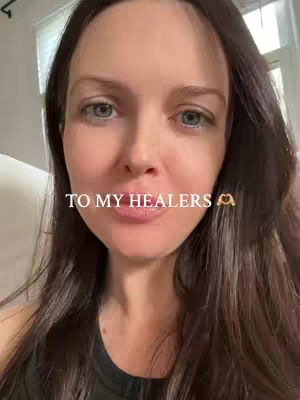A close-up shot of a woman looking directly at the camera, with text overlayed. She has fair skin, long dark hair, and is wearing a simple black top. The text "TO MY HEALERS" appears in white font, accompanied by a small hand-heart emoji. The background appears to be an indoor setting, possibly a room with a window.