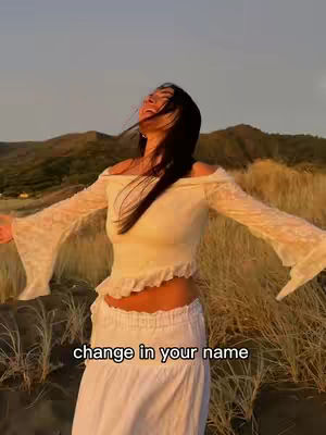 A young woman stands outdoors with arms outstretched, looking upwards with a serene expression. She wears a cream-colored, off-the-shoulder top and a long white skirt. The background shows a field of tall, dry grass and rolling hills. It is a bright day, and the lighting is warm, highlighting the subject with a soft glow. The image conveys a sense of peace and freedom.