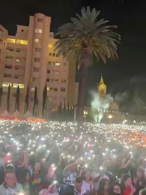 A night shot of a large crowd holding up illuminated phones, with a building and a palm tree in the background. A church stands in the distance.