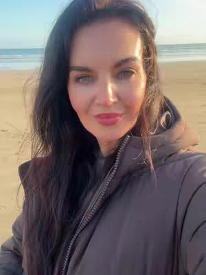 A selfie-style shot taken on a beach, with the creator centered in the frame, framed by an overcast sky. The creator is wearing a dark brown hooded coat. The background reveals a flat sandy beach and a seascape. The lighting suggests it may be sunset or sunrise.