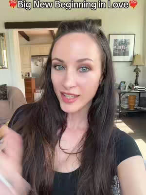 A woman with long brown hair is shown in a medium close-up, looking directly at the camera. She has fair skin, light eye makeup, and is wearing a black top. The background shows bits of a room. Above, text proclaims "Big New Beginning in Love" framed by heart emojis.