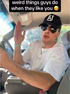 A man wearing a black cap and sunglasses is sitting in the driver's seat of a car, gesturing with his hand. The image is framed from inside the car.