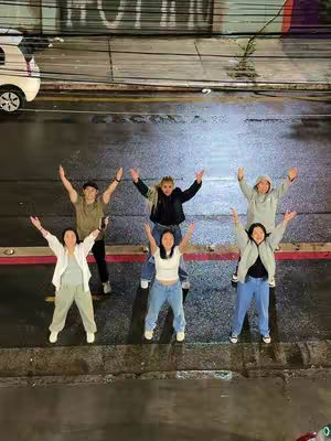 A group of six young people stand on a dark, wet street or pavement, arms raised in a celebratory pose. The image is taken from a high angle, looking down on the group, with the foreground dominated by their figures. The background shows indistinct city elements, possibly cars and buildings, with muted lighting and reflections.