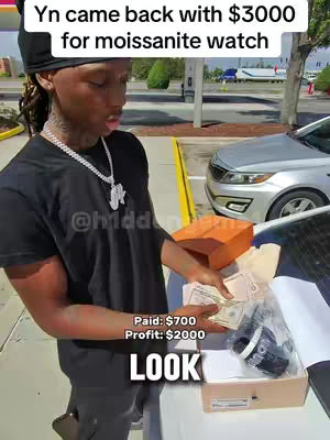 A young man stands on a parking lot with a black t-shirt, holding cash and a watch box near a car in front of a clear sky and building.