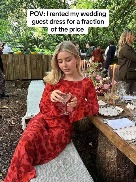 A woman in a red floral dress holding a drink at an outdoor wedding setting. The background features greenery, a wooden table set with dinnerware and candles. Text overlayed reads 'POV: I rented my wedding guest dress for a fraction of the price.'