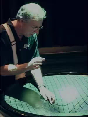 A man in a dark green shirt and glasses is bent slightly over a round, black-rimmed surface. The surface is a light green color, and has a grid pattern, with the middle portion curving downwards, as if something were pressing in on it. The man has his hand above the surface, as if he is about to touch it, with the finger pointing directly toward the center of the indented area. The background is completely dark.