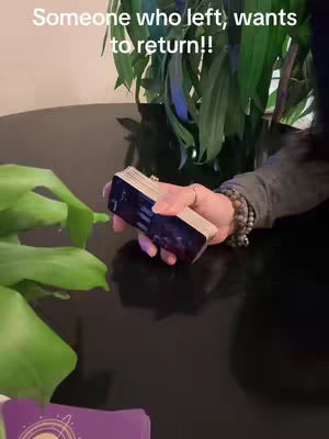A close-up shot of a person's hand holding a deck of tarot cards. The hand is adorned with bracelets and rings. The background features greenery and a dark, reflective surface.