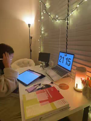 A cozy study setup with a person, likely a student, working on a tablet. The room is softly lit, with a fairy-light-decorated window in the background, enhancing the focus and productivity theme.