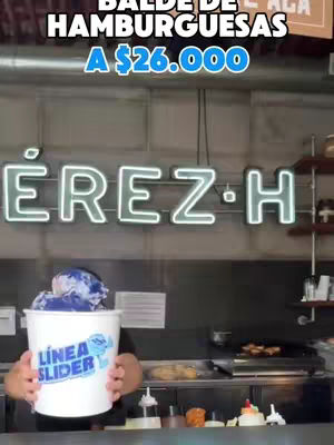 A person holds a white bucket filled with paper, with the words 'Linea Slider' in blue. Above, the name 'EREZ-H' is lit up in neon green. At the top, text announces the item as being 'BALDE DE HAMBURGUESAS...A $26.000'. The person stands behind a counter with cooking appliances and food items visible in the background.