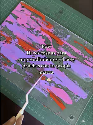 An overhead shot shows a rectangular acrylic board, diagonally positioned. The board is covered in a vibrant painting of overlapping strokes in shades of pink, purple, and orange against a green backdrop of a cutting mat with grid lines and numbers. A white tool partially overlaps the board in the bottom left corner. Text is layered over the artwork.