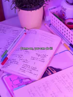 The image shows a desk scene with several scattered items. The scene centers on an open notebook filled with handwritten notes, positioned at the center. Several colorful pens are placed near the notebook, with another notebook partially visible beneath a pink planner. A small potted plant is situated at the top. The background is softly illuminated by purple lighting.