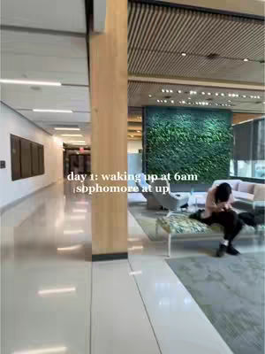 The image shows a well-lit hallway with a person sitting on a bench. The hallway has a glossy floor, and a wall on the left with some framed artwork. The right side has a wooden pillar and a wall decorated with a green plant panel.
