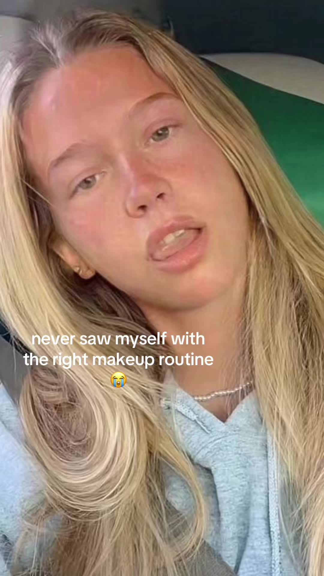 A selfie of a young woman is shown from the chest up, with her face taking up most of the frame. She has long, light blonde hair, and her skin is fair. She's wearing a light blue hoodie. The background appears to be the interior of a vehicle, with a green stripe on the upper right. The hook text is overlaid on the bottom of the image containing the text 'never saw myself with the right makeup routine' and an emoji between the text and the routine word. 