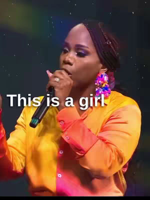 A woman with dark skin is holding a microphone and singing on a stage. The background is dark with some dim lights. She is wearing a yellow and orange blouse and large, colorful earrings. Text overlayed on the image reads 'This is a girl'.
