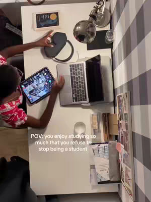 An overhead shot of a workspace with a person working at. The person, with visible arms and a red shirt with white patterns, is seated at a white desk. On the desk are several items: a laptop, an iPad with a colorful display, headphones, a lamp, and various stationary. The background includes a striped wall. The text overlay reads, 'POV: you enjoy studying so much that you refuse to stop being a student.'