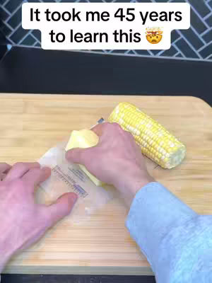 A close-up shot of hands preparing corn on the cob. The scene is a wooden cutting board with a partially shucked corn cob and a stick of butter being unwrapped. The text overlay reads, 'It took me 45 years to learn this'. The background is a black and white tiled texture.