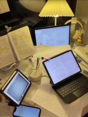 An overhead shot of a desk setup for studying is shown, with notebooks, papers, and electronic devices. A laptop, a tablet with a keyboard, and a smartphone are visible, all displaying lit screens. The scene is illuminated by a warm, yellow-toned desk lamp. A glass with a spoon and a small bottle with some liquid sits on the desk.