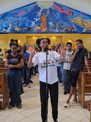 A photo taken inside a church. A young woman stands in the center, facing the camera, with her arms raised and hands open as if gesturing. She is wearing a white t-shirt and black pants and is speaking into a microphone. Behind her are a group of seated people. The background shows the interior of a church.