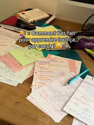 A close-up shot of a wooden desk covered in papers, notes, and a few books. The papers appear to be filled with handwritten notes, and various colored sticky notes are scattered among them. In the foreground, a blue pen rests on top of the documents, suggesting an active study or note-taking session. Behind the papers, there's a glimpse of a computer screen, adding a digital element to the otherwise analog workspace. The overall composition gives a sense of study.