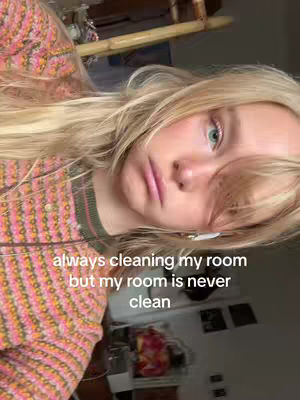A person with long blonde hair is shown in a selfie-style shot, angled with their upper body and face. The person is wearing what seems to be a patterned sweater with pink and orange tones. The background is out of focus, revealing hints of a room setting. Overlayed text at the bottom of the image states 'always cleaning my room but my room is never clean'