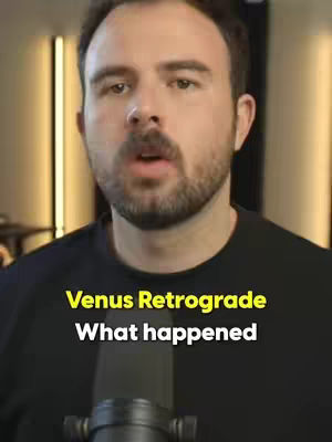 A medium shot of a man indoors, facing the camera. The man has short, dark hair and a beard. He wears a black shirt and speaks into a black microphone. Overlayed on the image is text in a yellow font, which reads, "Venus Retrograde, What happened". The background appears to be a plain wall with some indication of lights.