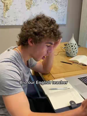 A young man with curly brown hair is sitting at a wooden table, looking down with a stressed expression. He's wearing a gray t-shirt and a silver chain. There's a laptop and a notebook open in front of him. In the background, there's a world map and a decorative vase.