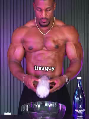 A muscular man, shirtless, stands in front of a light purple/pink vertically striped background. He holds an icy, cloudy object in his hands in front of a glass bowl. A bottle of water sits on the right.