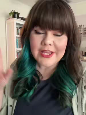 A woman with long brown with the green-colored hair is speaking to the camera with her right hand slightly raised. She wears a dark top and a light green jacket. The background is a room with a white cabinet with books in it and white wall, and a small plant is on top of the cabinet.