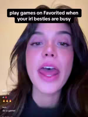 A selfie-style video frame featuring a woman with dark hair and a black top. She's positioned close to the camera, with her mouth open, exposing her tongue. A text overlay in a black box reads, 'play games on Favorited when your irl besties are busy.' The background is blurred and neutral, with a minimal color palette dominated by the woman's skin tones and pink lipstick.