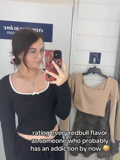 A young woman with dark hair and glasses is taking a selfie in front of a mirror in a clothing store dressing room. She's wearing a black top with a white trim and holding a phone with a floral case up to her face. Text overlayed on the image reads: 'rating every redbull flavor as someone who probably has an addiction by now'. A rack of clothes and a sign are visible in the background.