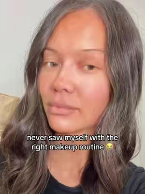 The image is a close-up of a woman's face with long, wavy, dark brown hair. The woman is looking slightly down and to the side. The background is a plain off-white. Text overlayed at the bottom says, 'never saw myself with the right makeup routine' and has a slightly sad face emoji. The lighting is soft and natural, creating a gentle and inviting mood.
