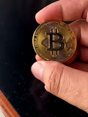 A hand holds up a gold Bitcoin coin against a blurred, dark background. The Bitcoin symbol is prominently visible. The hand and coin are the focus, with the rest of the image out of focus.