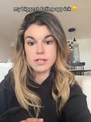 A selfie-style shot of a woman with long blonde hair, wearing a black hoodie, looking directly at the camera with a neutral expression. The background is a softly lit living space with a glimpse of a dining area. Text overlayed at the top reads 'my biggest dating app ick' with a thinking face emoji.