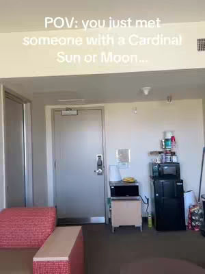POV shot of an apartment interior. The camera is pointed at a door. To the left, a fragment of a red upholstered seat is visible. Adjacent to the door, a small cabinet with a microwave is positioned, and on top of it, various bottles and containers are arranged.
