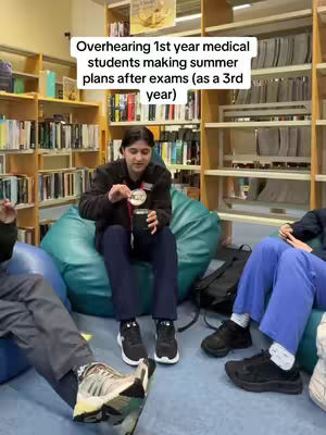 A person, presumably a student, sits on a large beanbag chair, holding a small black cup. The person is wearing a black jacket, a black shirt underneath, dark trousers, and sneakers. A light-colored tag is visible. Behind the person is a softly lit library with wooden bookshelves filled with books. To the side are other people seated in a similar setting, one wearing blue scrubs. The image is a medium shot, eye-level, with text overlay near the top.