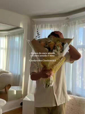 A person holds a bouquet of flowers in front of them. The person is in an indoor setting, possibly a home or apartment, with tall windows showing off the curtains. The overall color palette is soft and neutral, with light colors dominating the background. Text overlay is present.