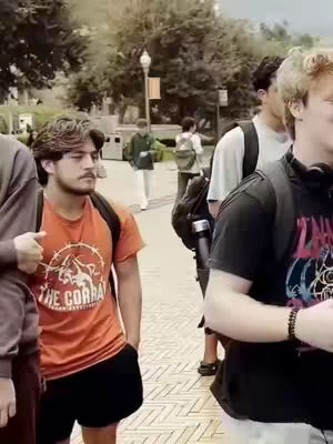 The image shows a group of young men walking on a paved pathway. The man in the foreground is wearing an orange t-shirt with an emblem and black shorts, while others in the background are carrying backpacks with a blurred background of buildings and trees.