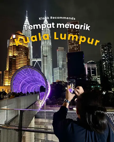 The cover image captures a nighttime cityscape featuring the Petronas Twin Towers illuminated in white, with an individual holding a phone taking a photo in the foreground. The urban landscape is completed with other tall buildings, and a bridge-like structure with purple lights extends into the composition, with text overlayed on top.