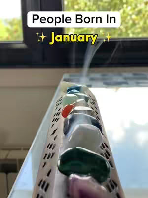 The image shows a close-up of a decorative display. In the foreground, there's a series of polished gemstones resting on a metal structure with a textured surface. Smoke is rising from the structure, adding a mystical element. The background is out of focus, with a window visible. The overall composition is centered, with the gemstones and smoke creating a vertical focal point.