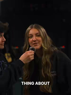 A woman with long, wavy blonde hair is centered in the frame, holding a microphone. To her left, the arm and shoulder of another person is visible, also holding a microphone. The image appears to be taken indoors with a dark background.