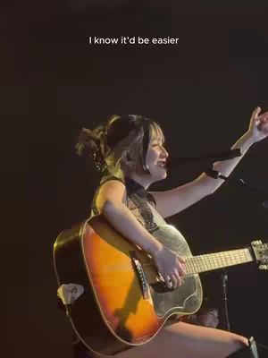 A woman is performing on stage, holding a guitar and singing into a microphone. She is wearing a black top and her hair is styled with light-colored highlights. The background is dark.